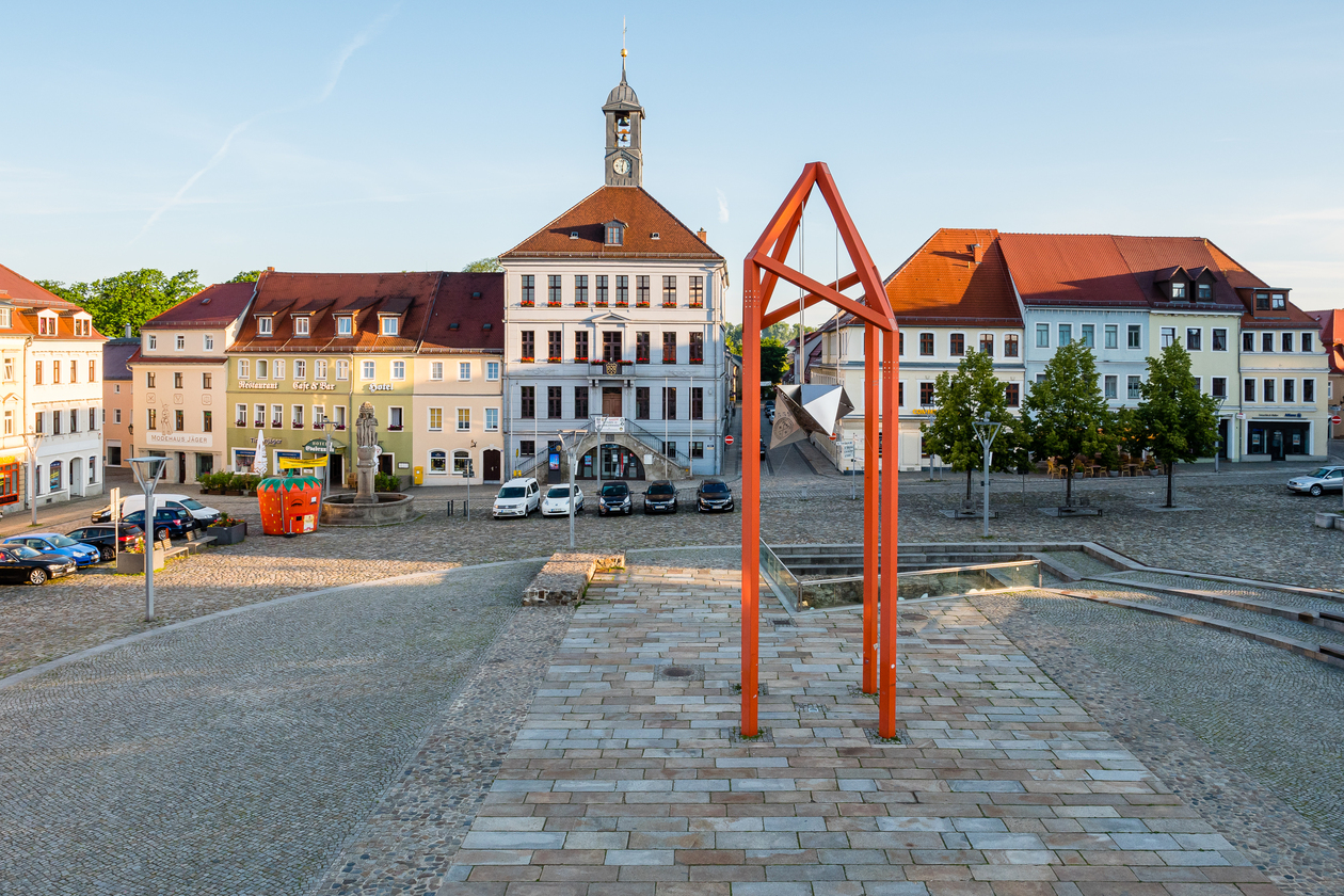 Das Foto zeigt ewine Marktplatz mitz Granitpflaster. Im Vordergrund ein Kunstwerk, bestehend aus Metallpfeilern. Sie deuten die Umrisse des Turmes des ehemaligen Rathauses an. Im Hintergrund eine Häuserzeile mit historishcen Fassaden und das Rathaus.