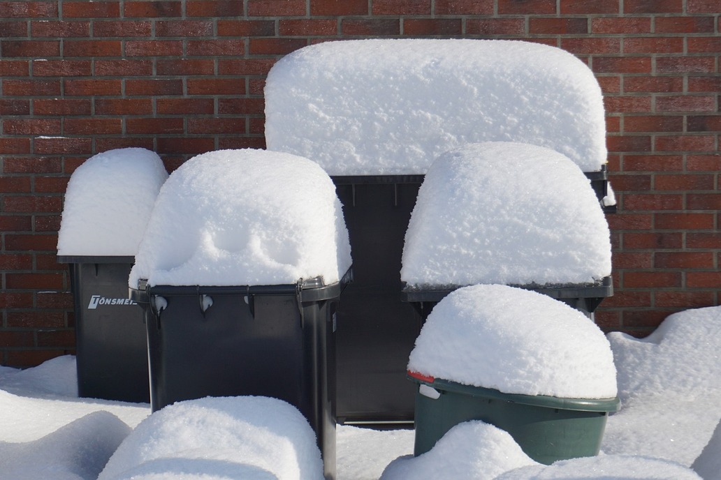 Vor einer Hausmauer stehen verschiedene tief verschneite Mülltonnen