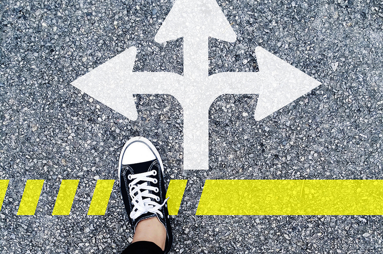 Drei Richtungspfeile auf einer Straße, vor den Pfeilen Füße in Turnschuhen