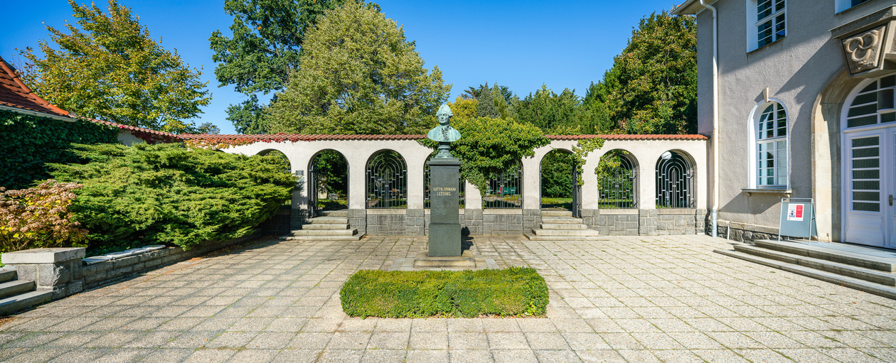 Das Foto zeigt einen Platz, in der Mitte eine Stele mit der Büste Lessings. Im Hintergrund ein Wandelgang,