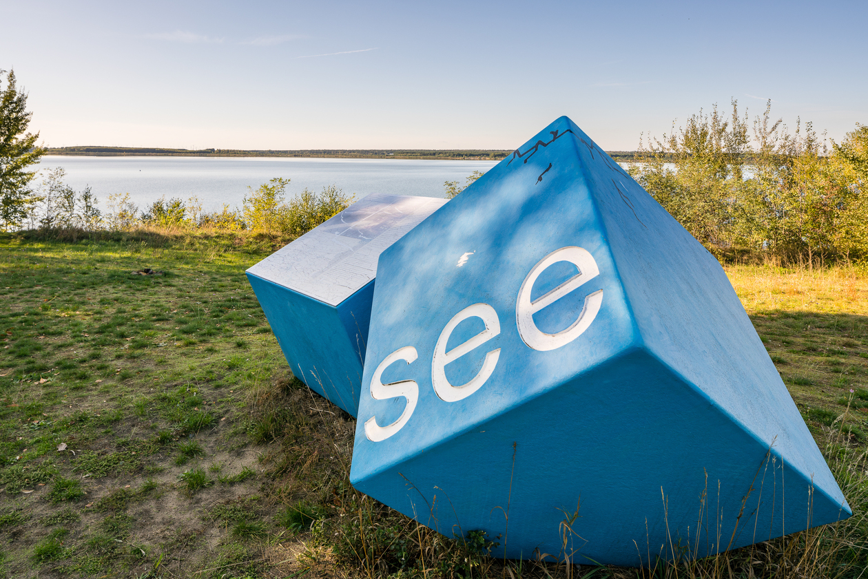 Das Foto zeigt einen See, am Ufer im Vordergrund zwei große blaue Würfel. Ein Würfel trägt den Schriftzug "see"