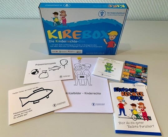 Das Bild zeigt den Inhalt der Kindergartenbox: verschiedene Medienträger, zwei Kinderbücher , eine Puppe und zwei Blechdosen