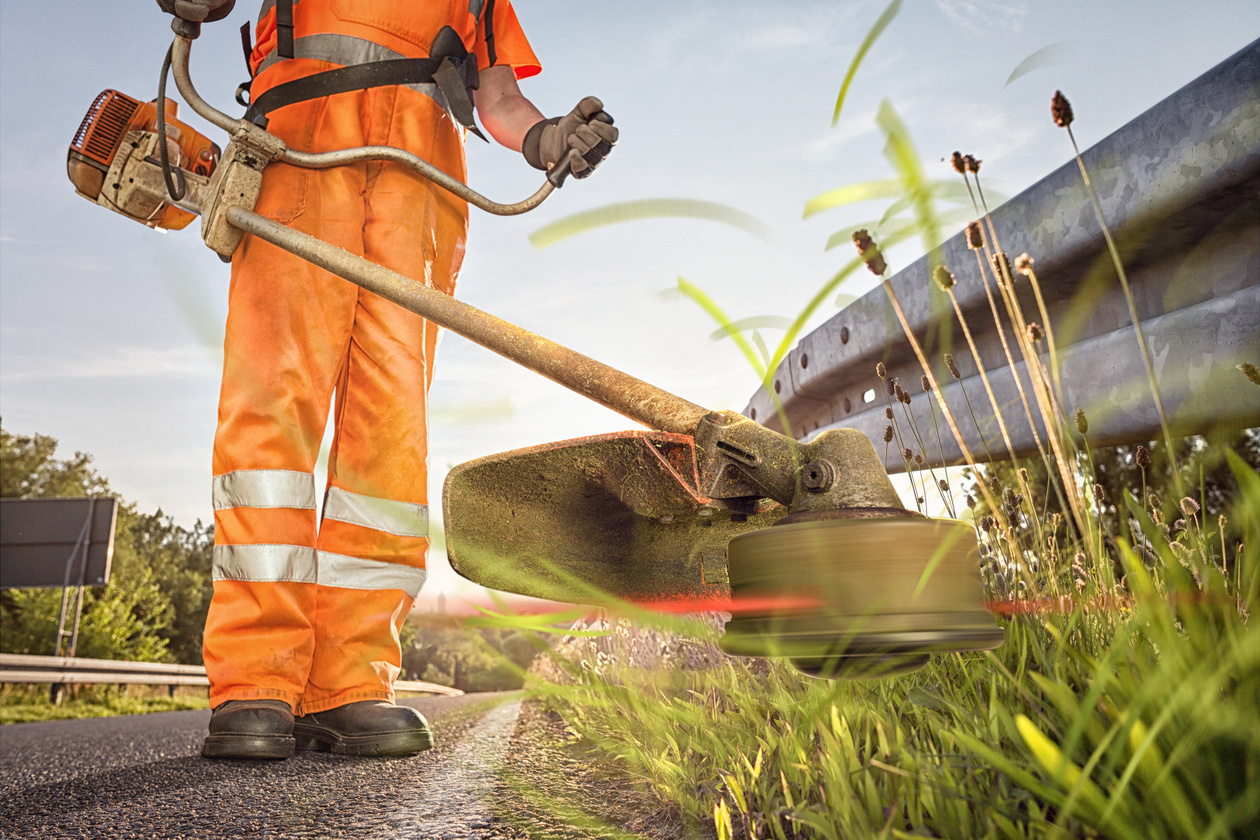 ein Straßenwärter in Arbeitskleidung mäht mit einem Arbeitsgerät vor einer Leitplanke