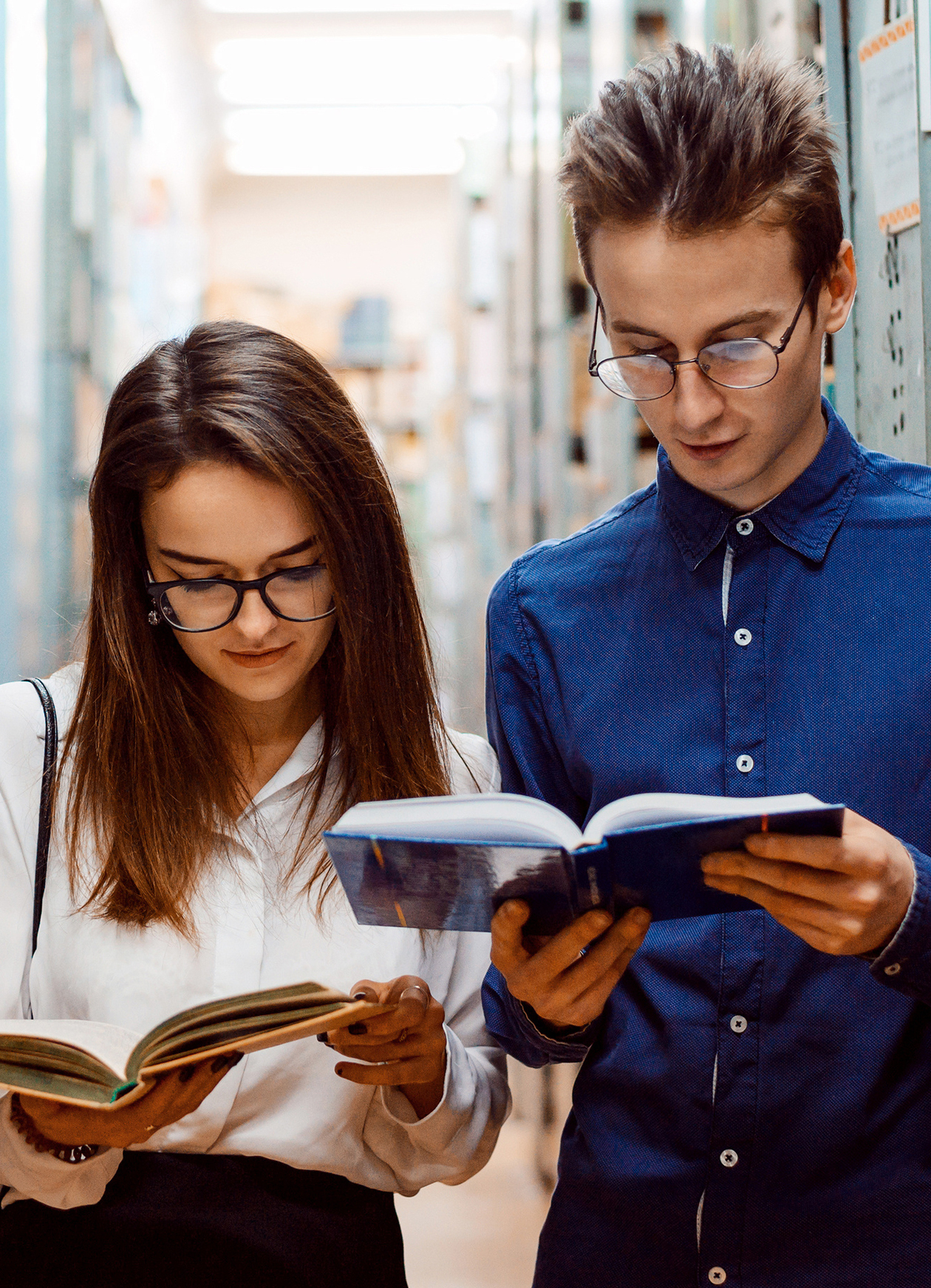 eine junge Frau und ein junger Mann stehen zwischen Regalen und lesen in Büchern