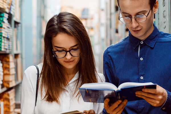 eine junge Frau und ein junger Mann stehen zwischen Regalen und schauen in ein Buch