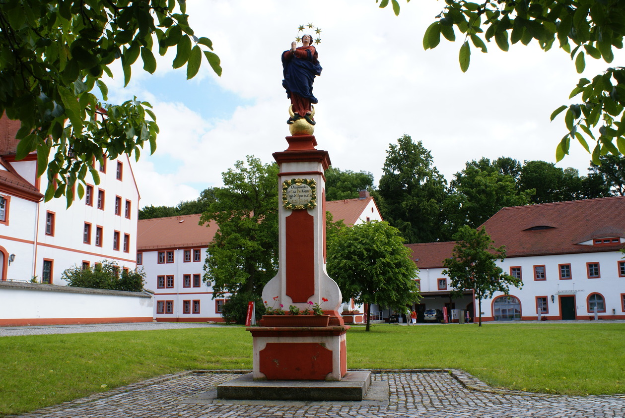 Klosteranlage: verschiedene Gebäude, Rasen, Bäume und im Vordergrund eine Dtele mit kirchlichem Heiligenmotiv.