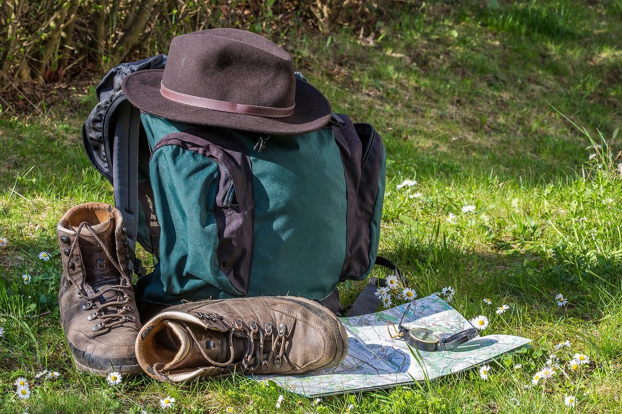 ein Rucksack, ein Hut und Wanderschuhe liegen auf einer Gänseblümchenwiese, daneben eine Karte und ein Kompass