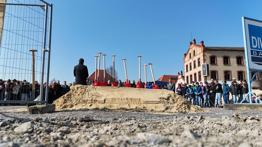 ein Sandhaufen mit acht Spaten, im Hintergrund viele Menschen