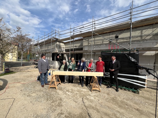 vor einem Rohbau mit Gerüst stehen 7 Menschen und schlagen Nägel in einen Balken