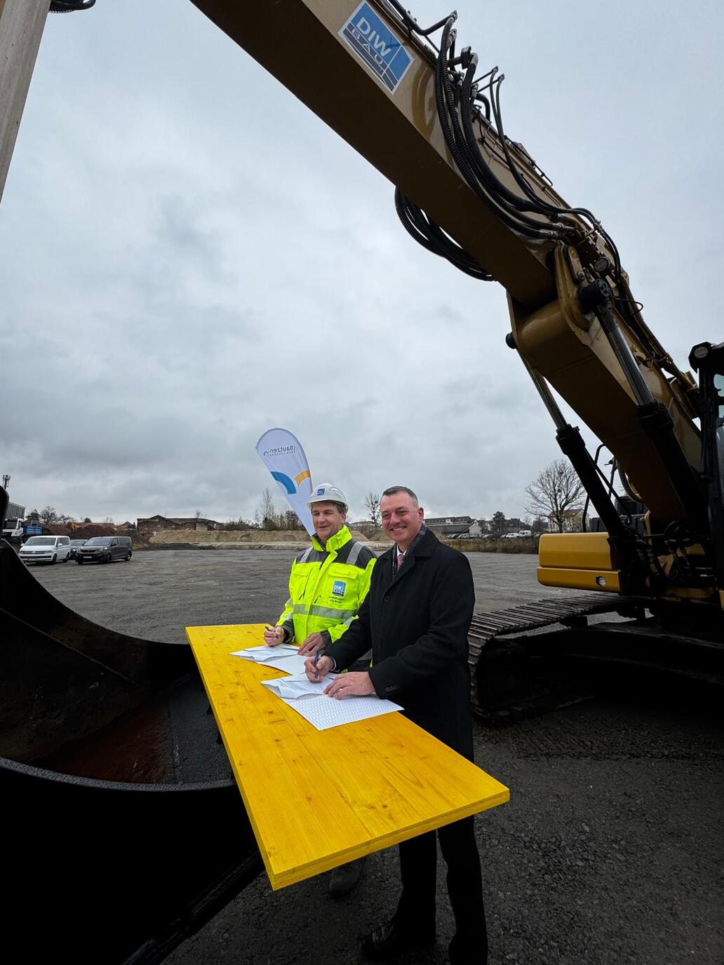 2 Personen unterzeichnen einen Vertrag, welcher auf einer Baggerschaufel liegt 