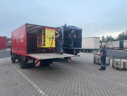 Ein LKW der feuerwehr und einer des technischen Hilfswerkes stehen auf einem Parkplatz. Drei Männer verladen Container