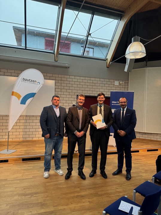 in einer Halle stehen vier Männer. Der dritte hält eine Mappe