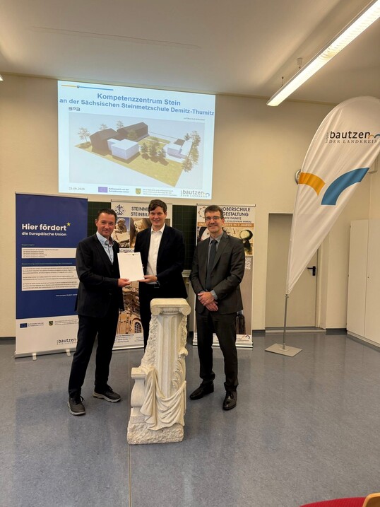 Drei Männer stehen hinter einem Steinsockel und halten ein Dokument in die Kamera, im Hintergrund eine Präsentation