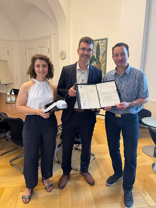 Eine Frau und zwei Männer halten eine Mappe mit Dokumenten in die Kamera. Die Frau hält eine VR-Brille.