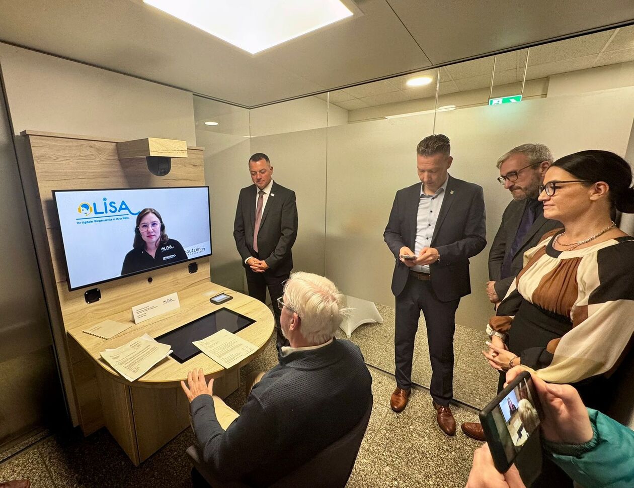Vor einem Tisch mit einem Bildschirm sitzt ein Mann. Auf dem Bildschirm ist eine Frau zu sehen und das Logo LISA. im Hintergrund stehen drei Männer und eine Frau und schauen zu.