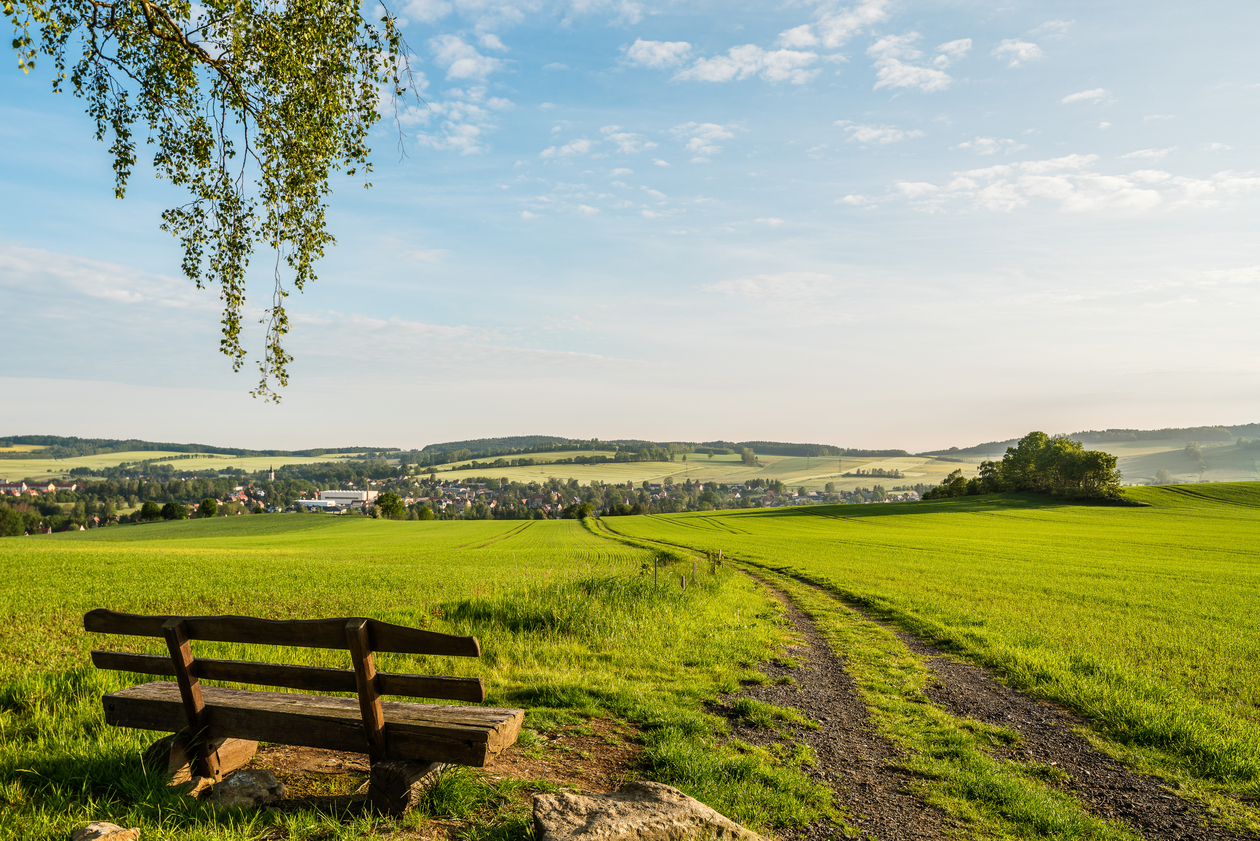 Eine Bank an einem Feldrand im Frühsommer mit Blick auf eine Siedlung und auf Hügelland