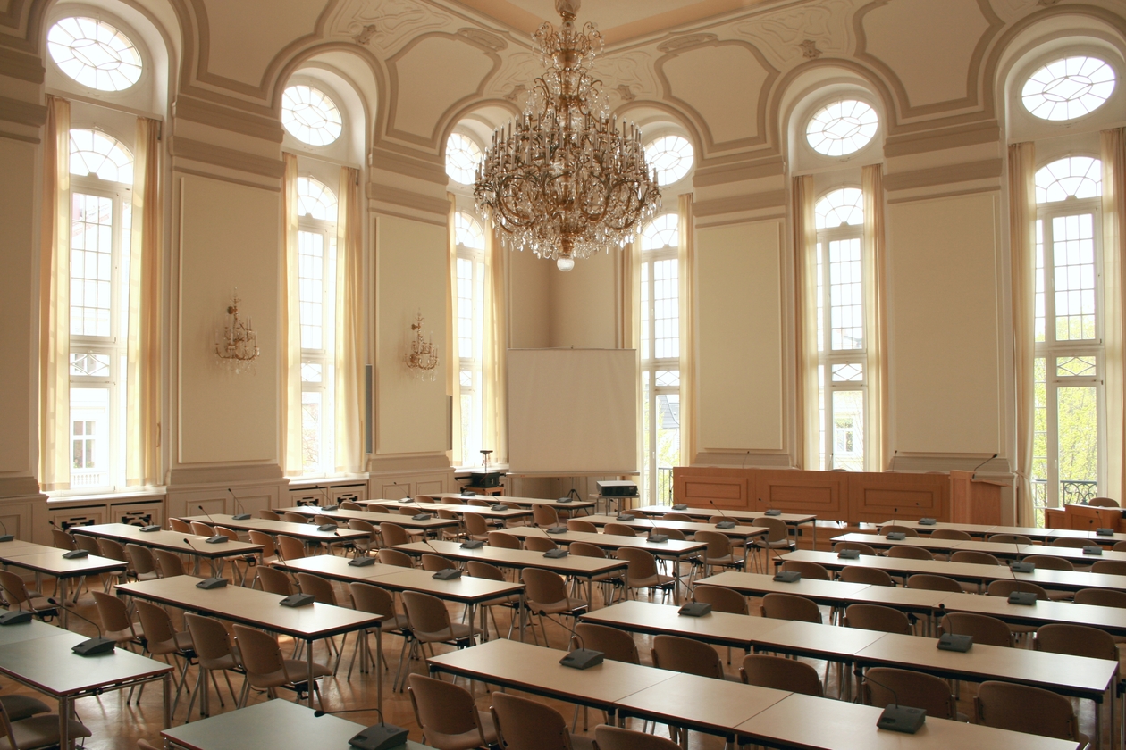 Ein Saal mit hohen Fenstern und einem Kronleuchter. Im Saal Bänke und Tische, die zu einer Sitzung aufgestellt wurden.