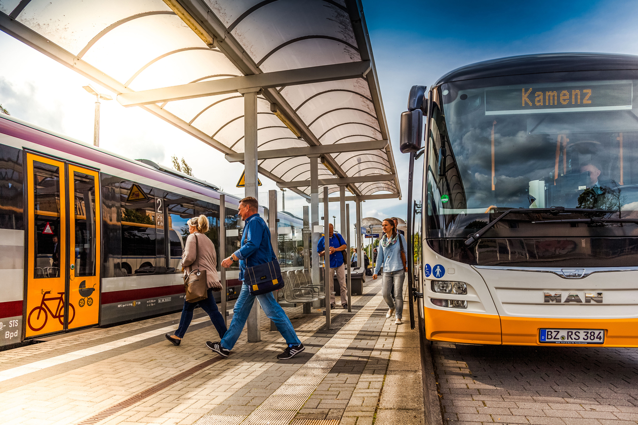 Eine Haltestelle mit Bus- und Bahnanbindung: Rechts ein Bus, links eine Bahn. Menschen, die umsteigen.