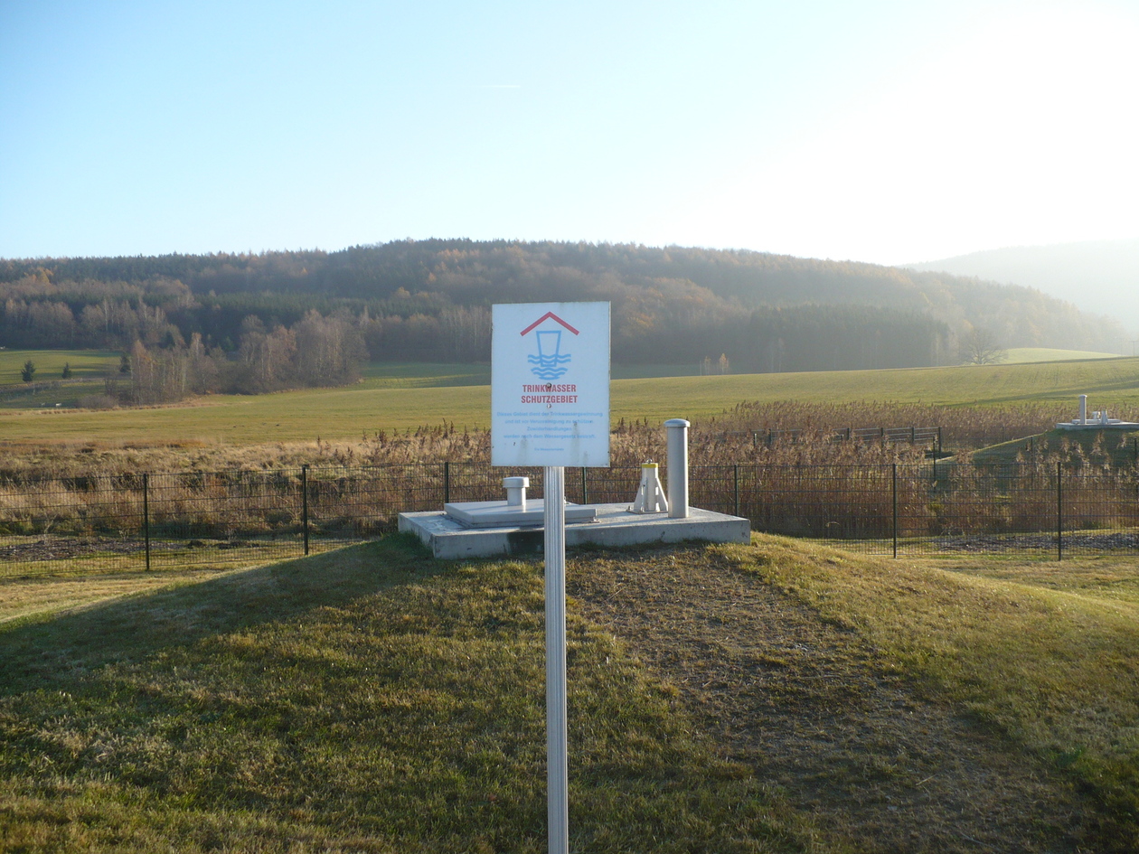 Das Bild zeigt eine Wasserfassung, davor ein Schild mit dem Hinweis auf das Trinkwasserschutzgebiet, im Hintergrund herbstlicher Wald.