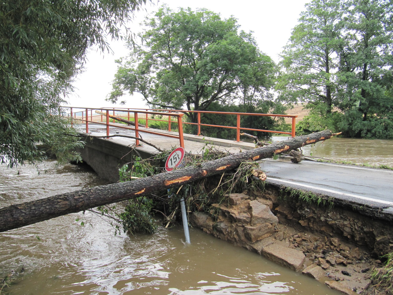 Auf dem Bild ist eine Brücke über einem Fluss zu sehen. Der Fluss ist über die Ufer getreten und hat Teile des Brückenbauwerks zerstört. Ein Baum ist umgestürzt und versperrt die Straße über der Brücke.