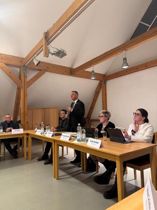 Zwei Frauen und zwei Männer sitzen an einem Tisch, ein Mann steht und spricht.