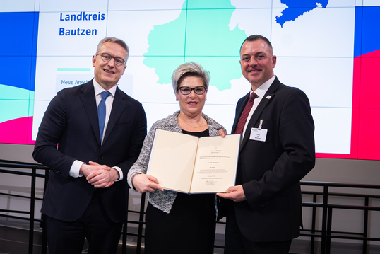 Zwei Männer und eine Frau halten eine Mappe mit Dokumenten in die Kamera