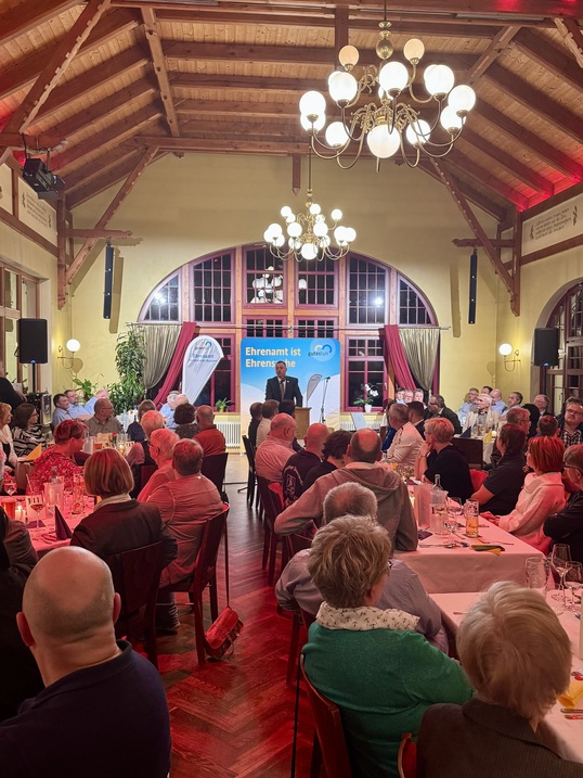 Sie sehen viele Menschen in einem großen Saal. In der Mitte steht Landrat und spricht zu den Gästen.