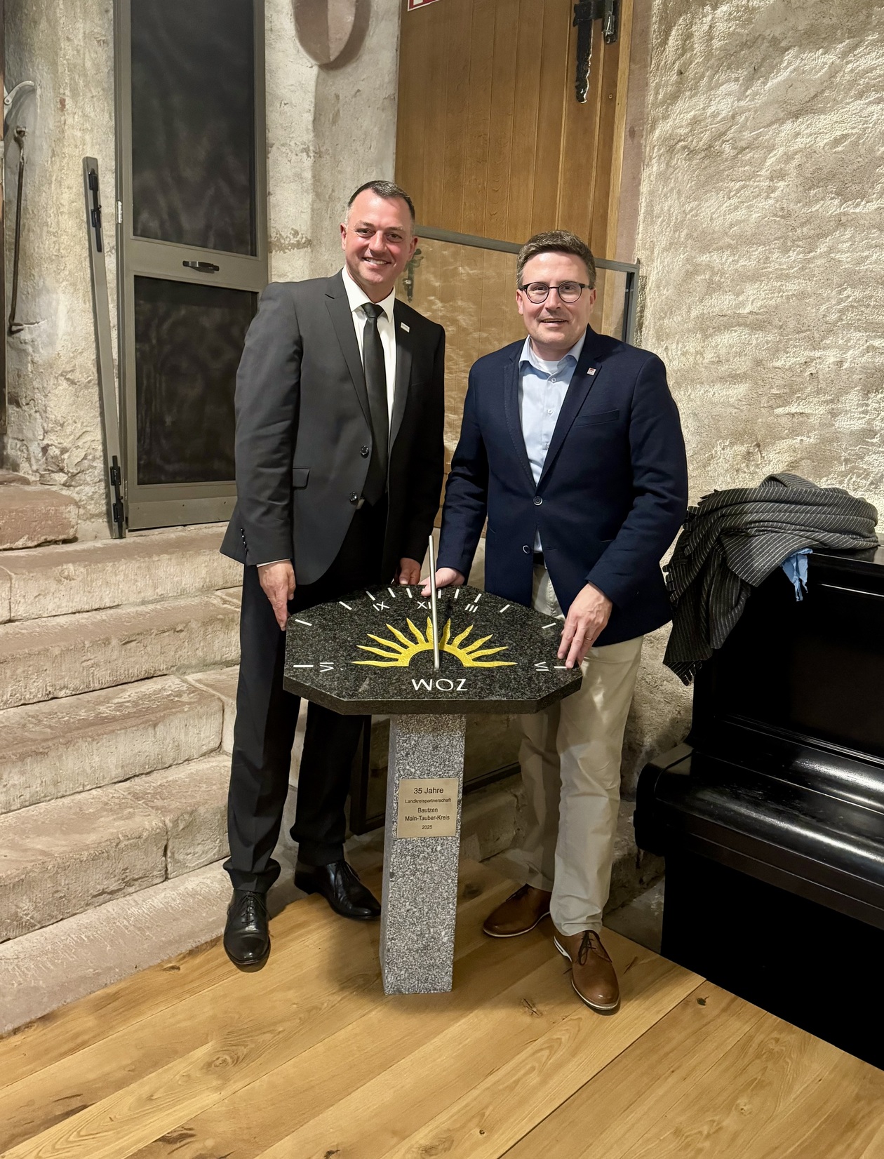 Sie sehen den Landrat von Bautzen links und den Landrat von Main-Tauber-Kreis rechts stehend. In der Mitte eine Sonnenuhr als Geschenk.