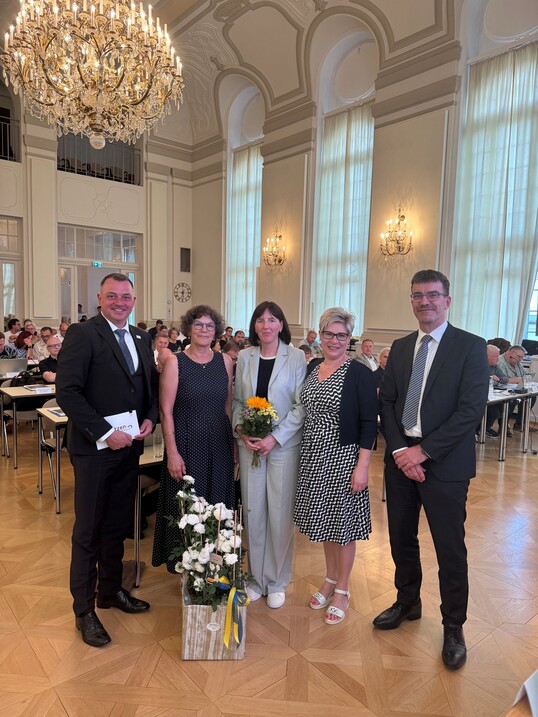 zwei Männer und drei Frauen schauen in dei Kamera, vor ihnen steht ein Rosenstock